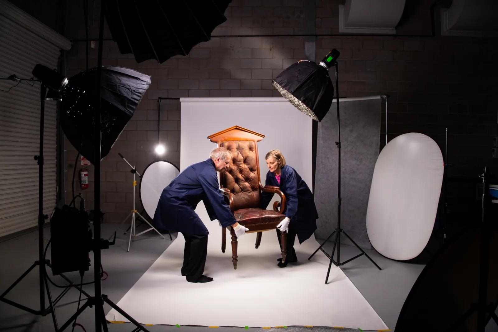Two people in blue protective smocks and white gloves carefully moving a vintage upholstered armchair on a white backdrop, surrounded by professional photography studio lighting and equipment in Sovereign Hill