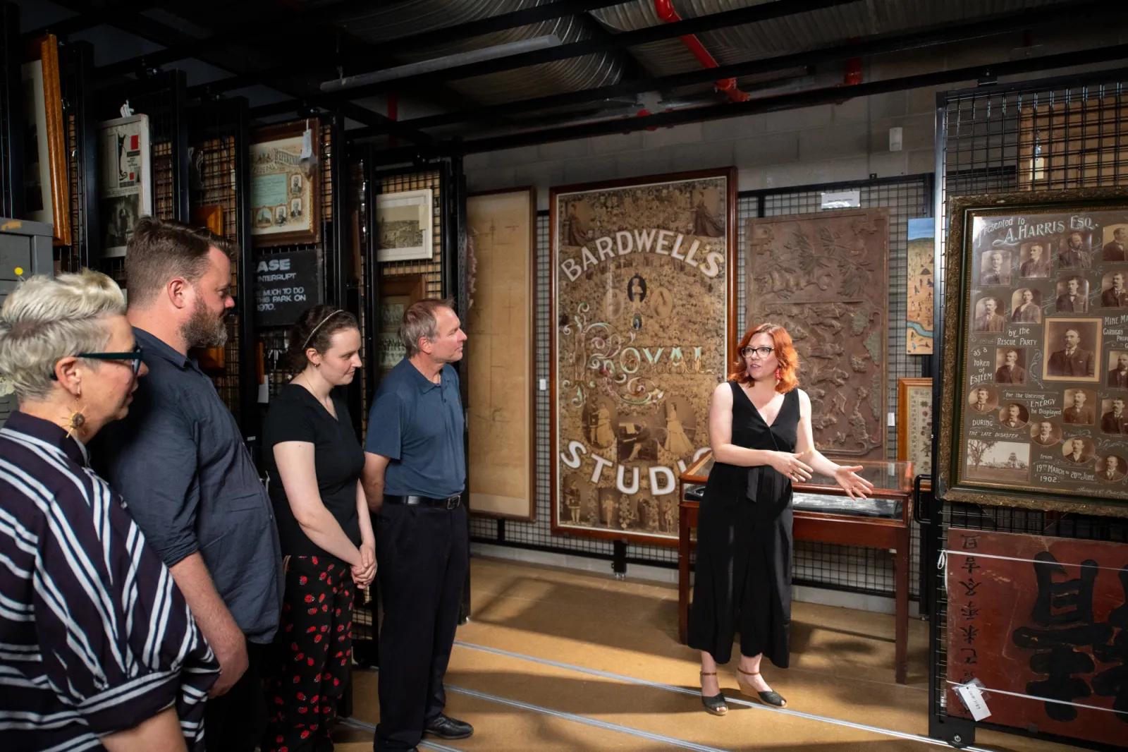 An expert leading a tour through an archive or historical exhibit in Sovereign Hill, pointing out details on large framed historical advertisements and portraits to a small audience.