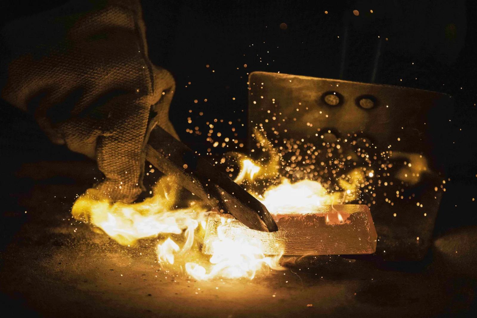 Close-up of a metal piece being forged, with sparks and flames flying as a gloved hand uses a tool to shape it.