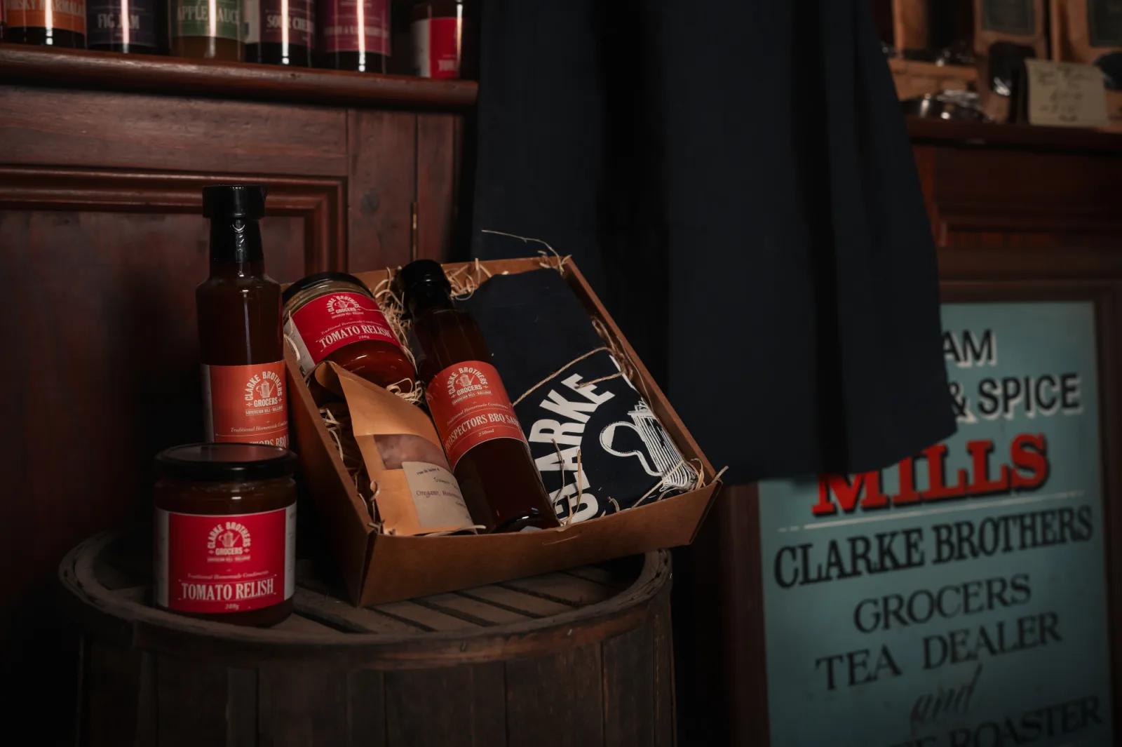 A rustic still-life photograph featuring a gift basket on a wooden barrel. The basket contains bottles and jars labeled "Tomato Relish" and "Clarke Brothers," along with a dark fabric item. The background includes a wooden wall with shelves holding various bottles and jars