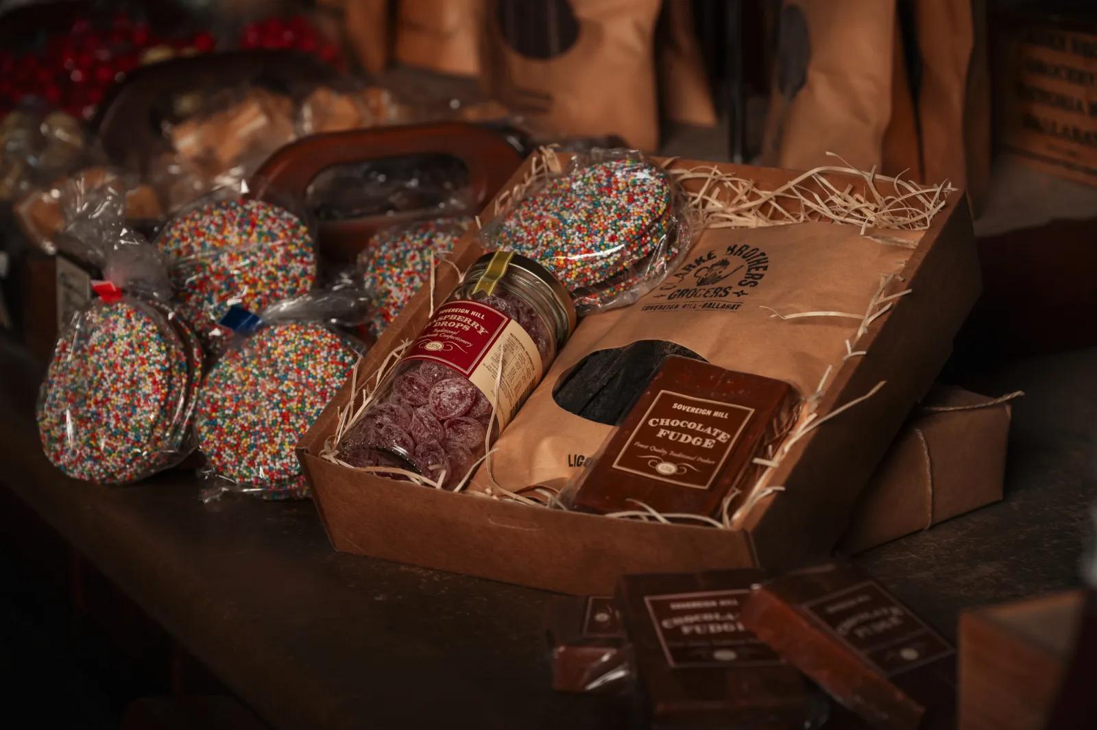 A close-up photograph of various food items packaged in a rustic style, including chocolate fudge, cookies, and preserves, possibly from a specialty food shop or market stall.