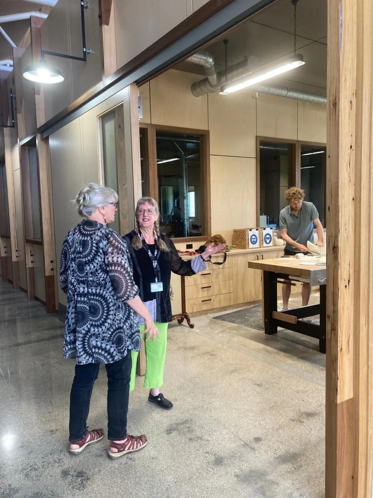 Two women standing in a workshop or studio, with one woman holding an object and gesturing, while a man works at a table in the background.
