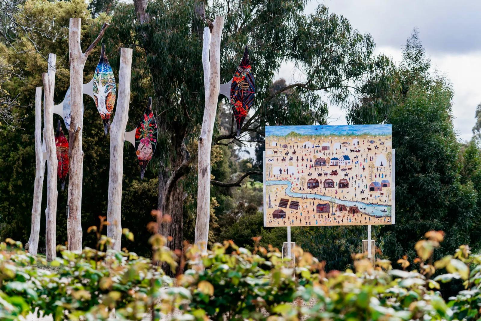 Outdoor art installation featuring three colorful glass lanterns hanging from white, branch-like structures and a square painting depicting a village or landscape on a clear stand, surrounded by greenery.