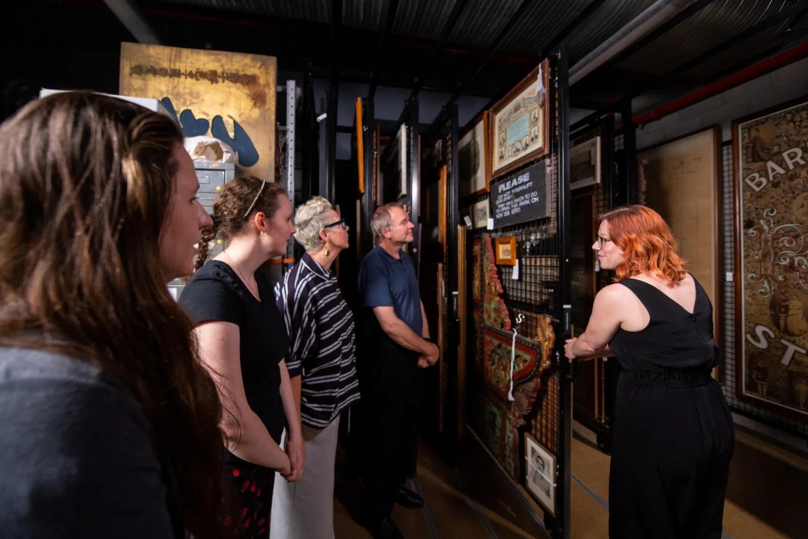 Group of five people looking at artwork in a gallery, including a woman in a black dress opening a sliding panel.