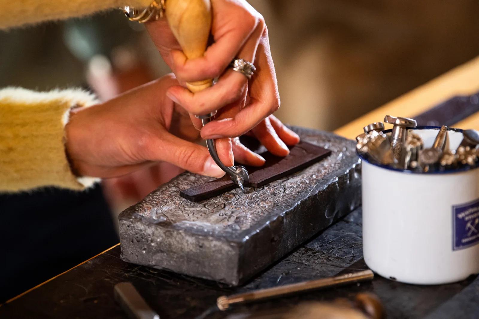 A close-up of hands using a metal stamping tool on a piece of leather placed on a metal block. The person wears rings and a yellow sweater. Nearby is a white container holding various metal stamping tools. The scene suggests leather crafting or metal engraving during the gold rush era in Ballarat