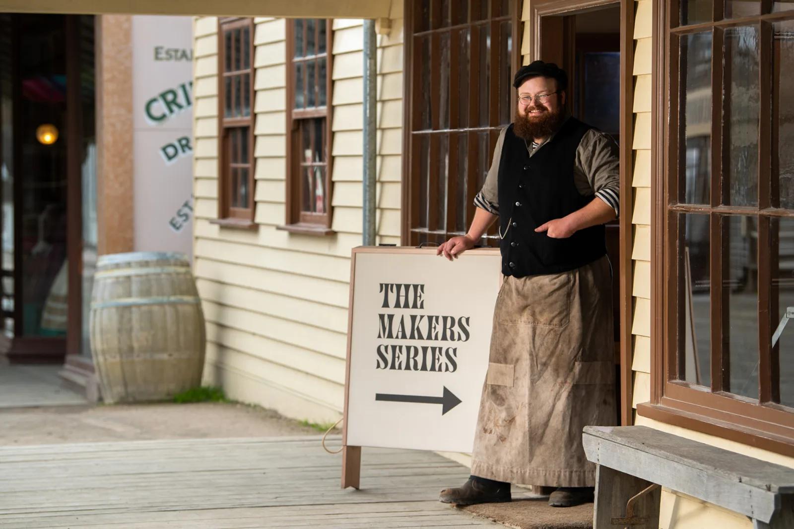 A man dressed in vintage attire, including a vest and long apron, stands in front of a wooden building, smiling. Beside him is a sign that reads THE MAKERS SERIES with an arrow pointing right. The setting resembles a historical or old Western town in Ballarat, with wooden sidewalks and a barrel visible in the background.