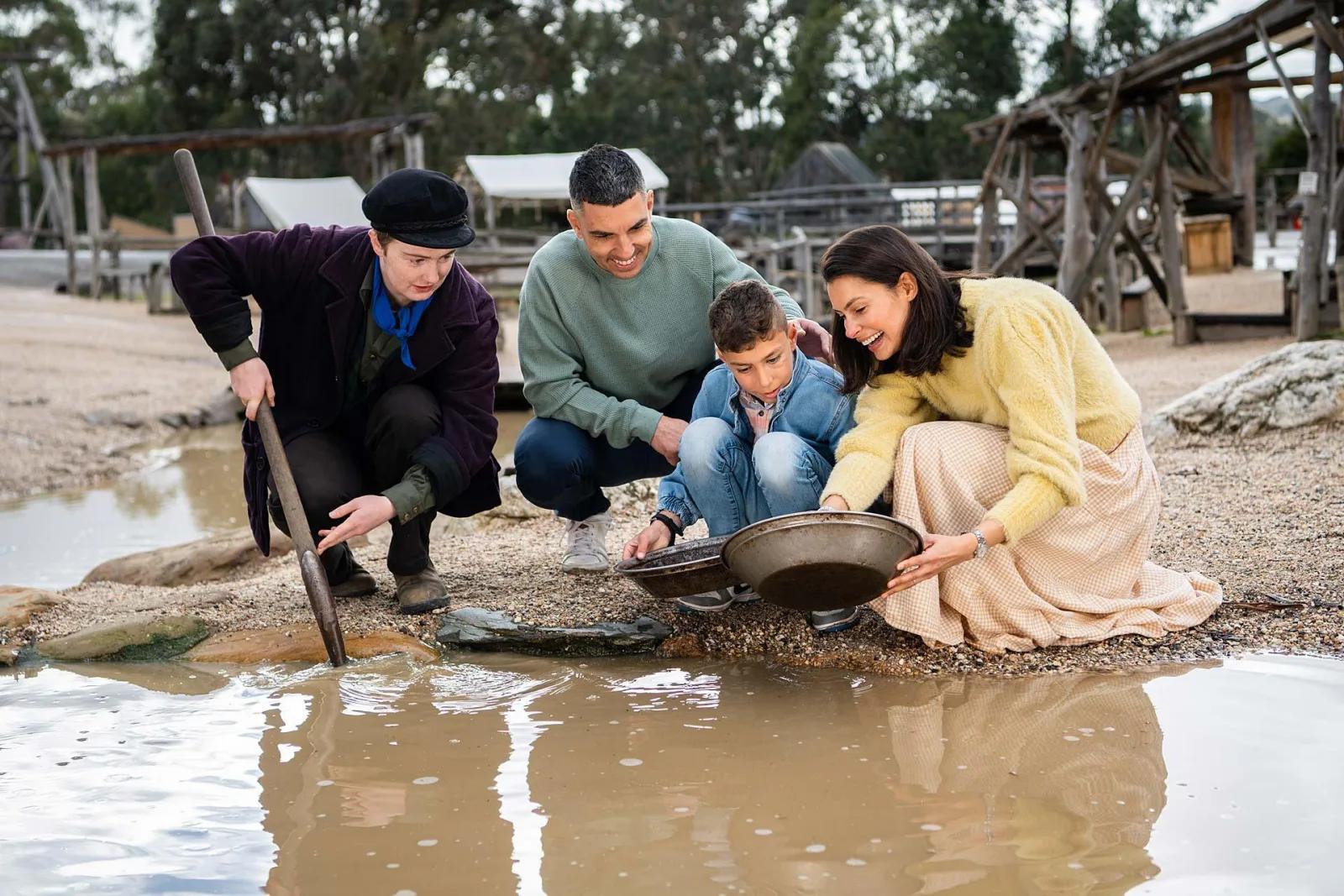 Two adults and two children are panning for gold at the edge of a muddy water pool in an outdoor setting. This is reminiscent of gold panning in Ballarat's gold rush era