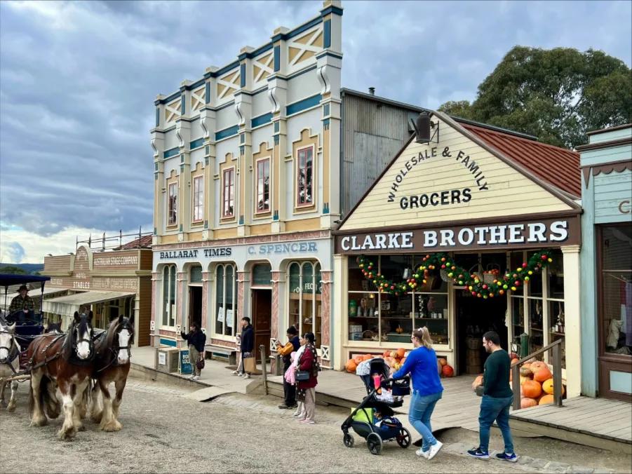 Visitors stroll along the historic main street at Sovereign Hill, where a horse-drawn carriage passes by iconic buildings including Clarke Brothers Wholesale & Family Grocers (adorned with festive garlands), Ballarat Times C. Spencer, and the ornate two-storey United States Hotel. The scene captures the lively, immersive atmosphere of this living gold rush museum.
