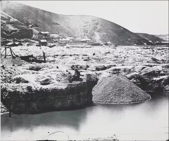 A historic black-and-white photograph depicts gold mining activity at Black Hill, with a miner panning in a water-filled sluice beside a large spoil heap; scattered tents and small buildings dot the barren, excavated landscape under a stark hillside, a scene preserved and interpreted at Sovereign Hill to illustrate the harsh realities of the Victorian gold rush.