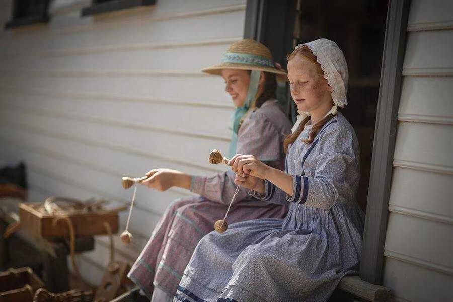 Two women in authentic 19th-century dresses sit on a porch at Sovereign Hill, gently spinning wool with drop spindles, capturing the quiet craftsmanship and daily life of the gold rush era, brought to life through immersive historical experiences at Sovereign Hill.