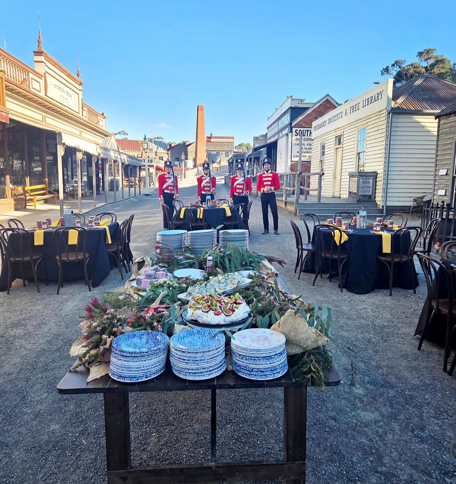 A meticulously arranged Main Street Function Setup at Sovereign Hill features a long wooden table adorned with fresh fruit, creamy desserts, floral arrangements, and stacks of vintage-style blue-and-white plates. Black-clothed dining tables with yellow napkins line the historic street, flanked by iconic buildings including the Criterion Store and Mechanics Institute & Free Library. Costumed performers in red military-style uniforms stand guard under a bright blue sky, capturing the immersive, celebratory atmosphere of event hosting within Sovereign Hill’s living gold rush town.