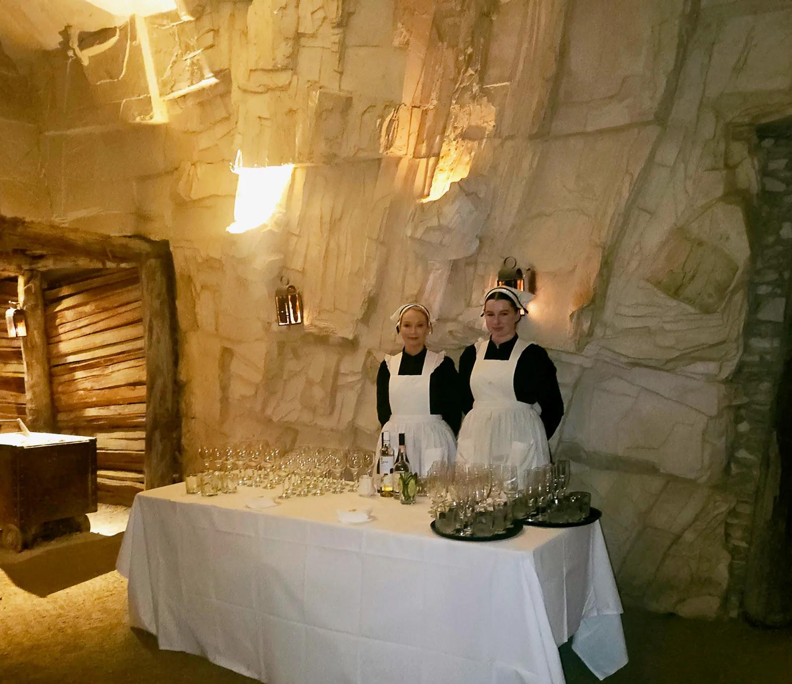 Two women in period-appropriate maid uniforms stand behind a white-clothed table set with glassware, bottles, and dining essentials inside the dimly lit, rustic stone chambers of Sovereign Hill’s Dining in the Mine experience. Warm lighting from vintage wall sconces illuminates the authentic underground setting, capturing the immersive historical atmosphere of this unique gold rush-era dining adventure at Sovereign Hill.