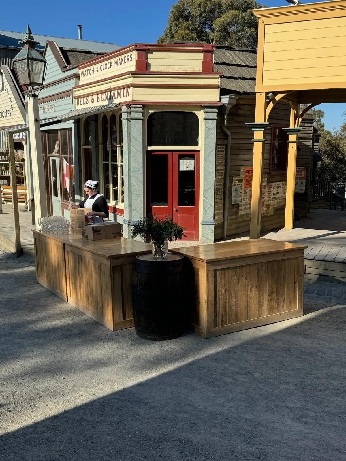A rustic Main Street bar setup at Sovereign Hill features wooden counters and a barrel, with a costumed interpreter working behind the counter near historic storefronts including Rees & Benjamin and Watch & Clock Makers. Sunlight bathes the scene, highlighting the immersive gold rush-era atmosphere of Sovereign Hill’s living history experience.