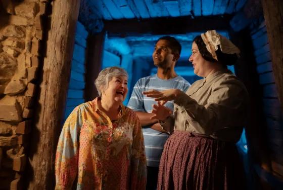 Three people stand inside a dimly lit, wooden tunnel with blue lighting. A woman in historical attire gestures while speaking to an older woman and a man in modern clothing, both smiling and engaged, suggesting a guided historical tour or interactive experience.